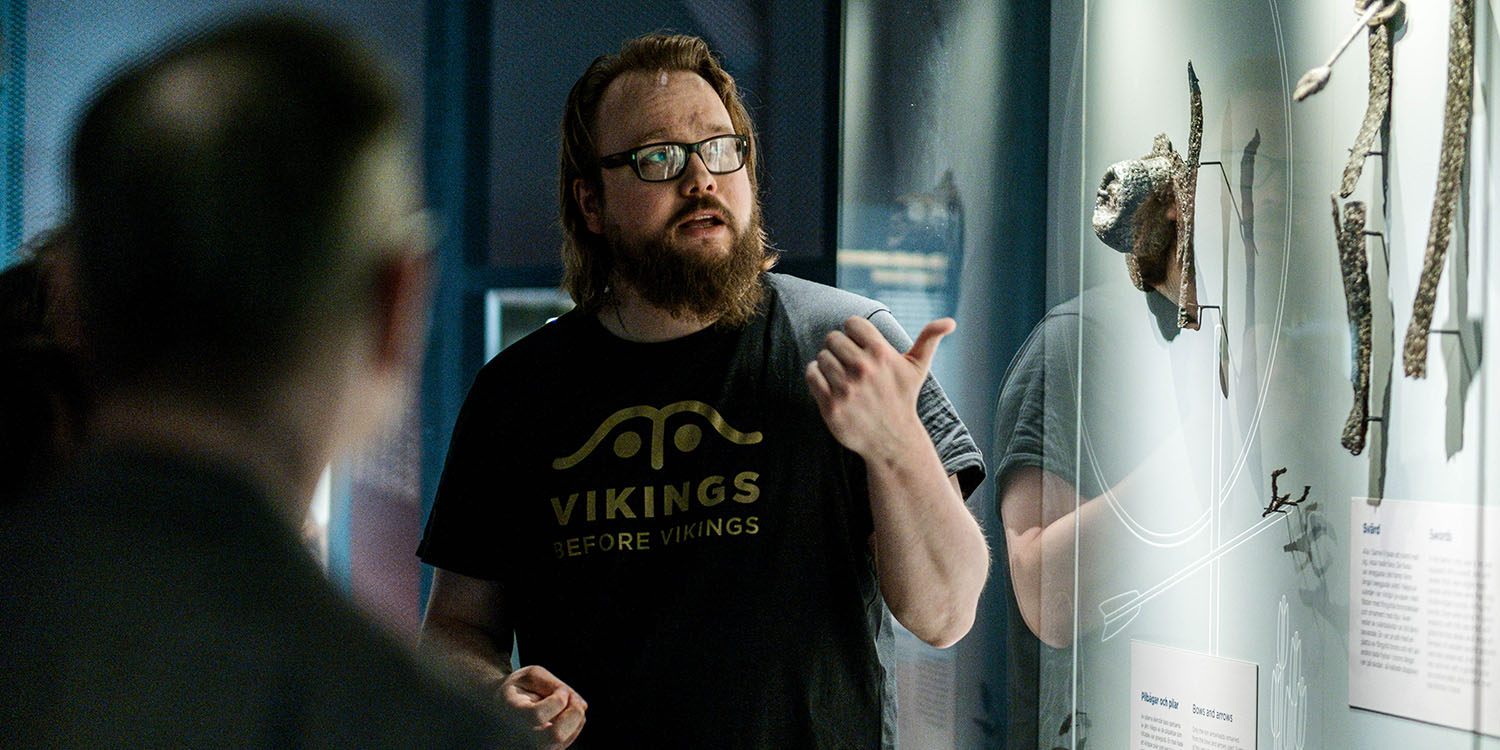 Guide med skägg och glasögon i svart tröja med tryck i guldfärg och texten vikings before vikings visar böjda svärd i en monter.